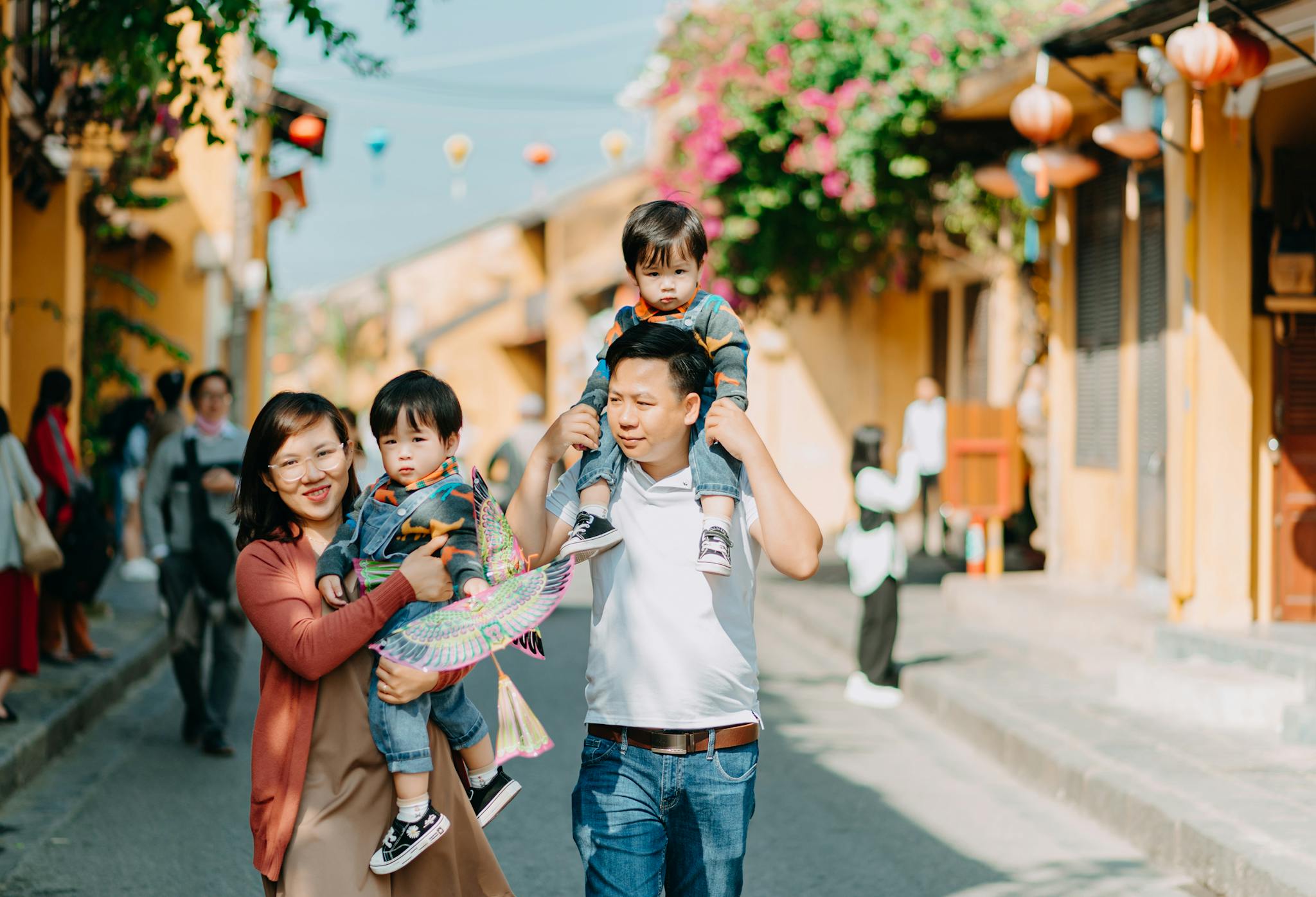 Cheerful Asian parents with sons walking together on asphalt road near buildings with hanging decorations on sunny street of town