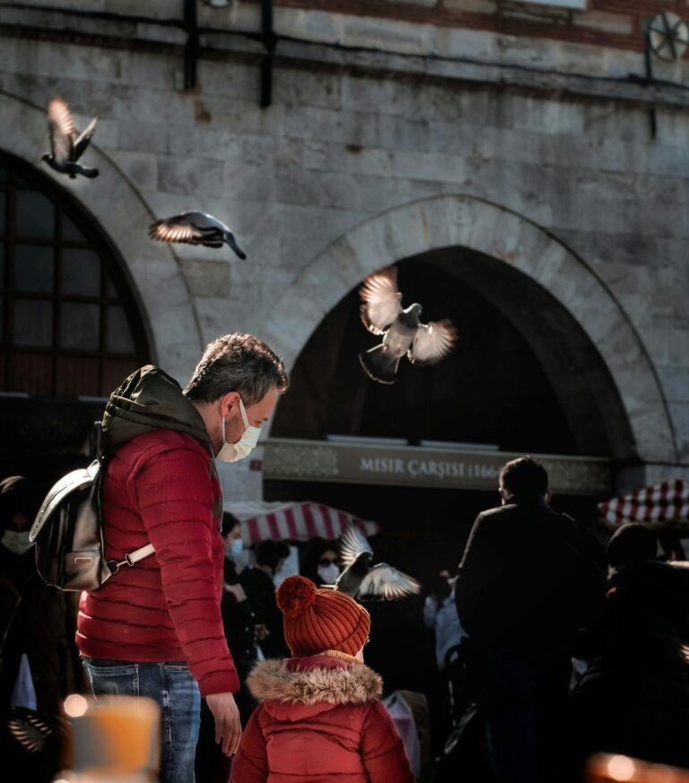 Father and son in red jackets watching flying pigeons in a lively market area.
