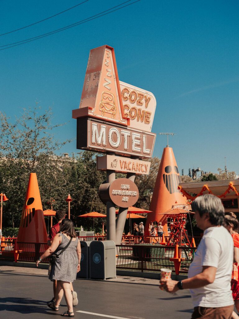 Vintage Cozy Cone Motel sign with people walking by in a lively amusement park setting.
